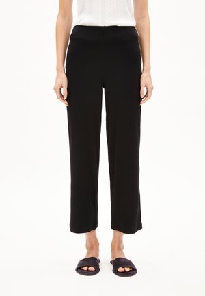 Mujer con pantalones negros de pierna recta, top blanco sin mangas y sandalias negras abiertas de punta, de pie sobre un fondo blanco.