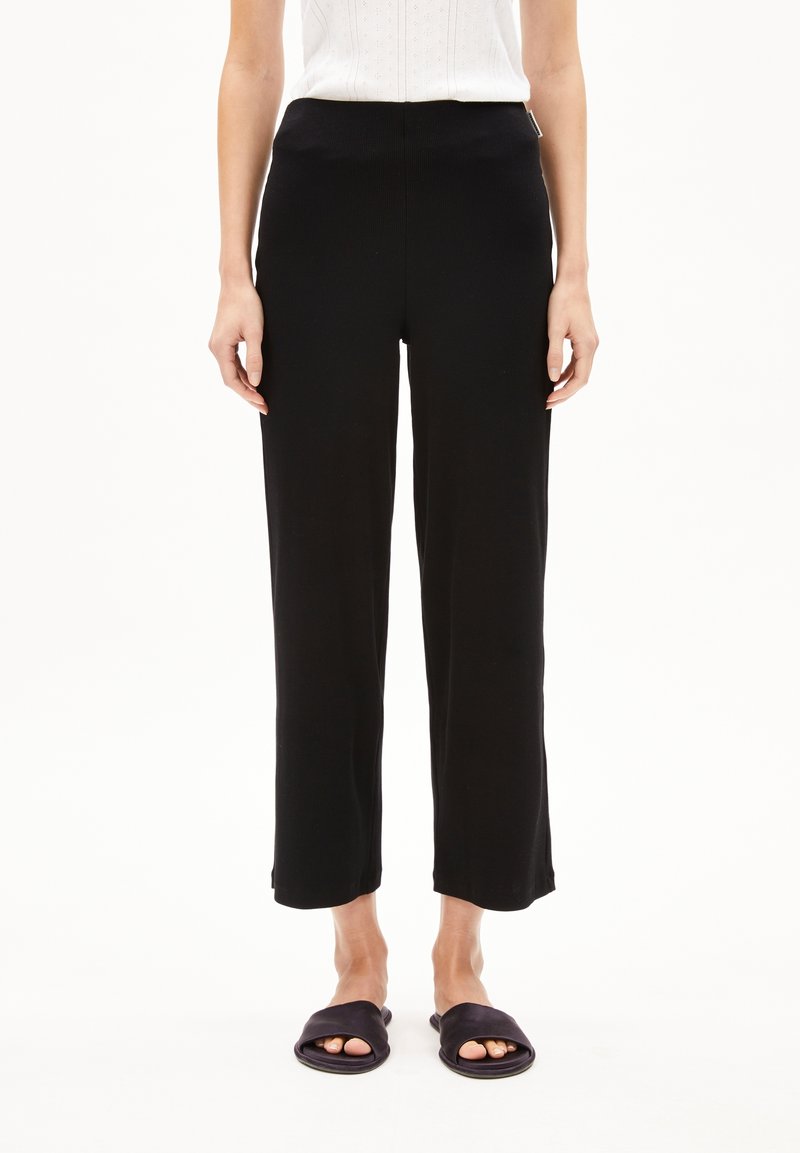 Femme portant un pantalon noir droit, un haut blanc sans manches et des sandales noires ouvertes, debout sur un fond blanc.