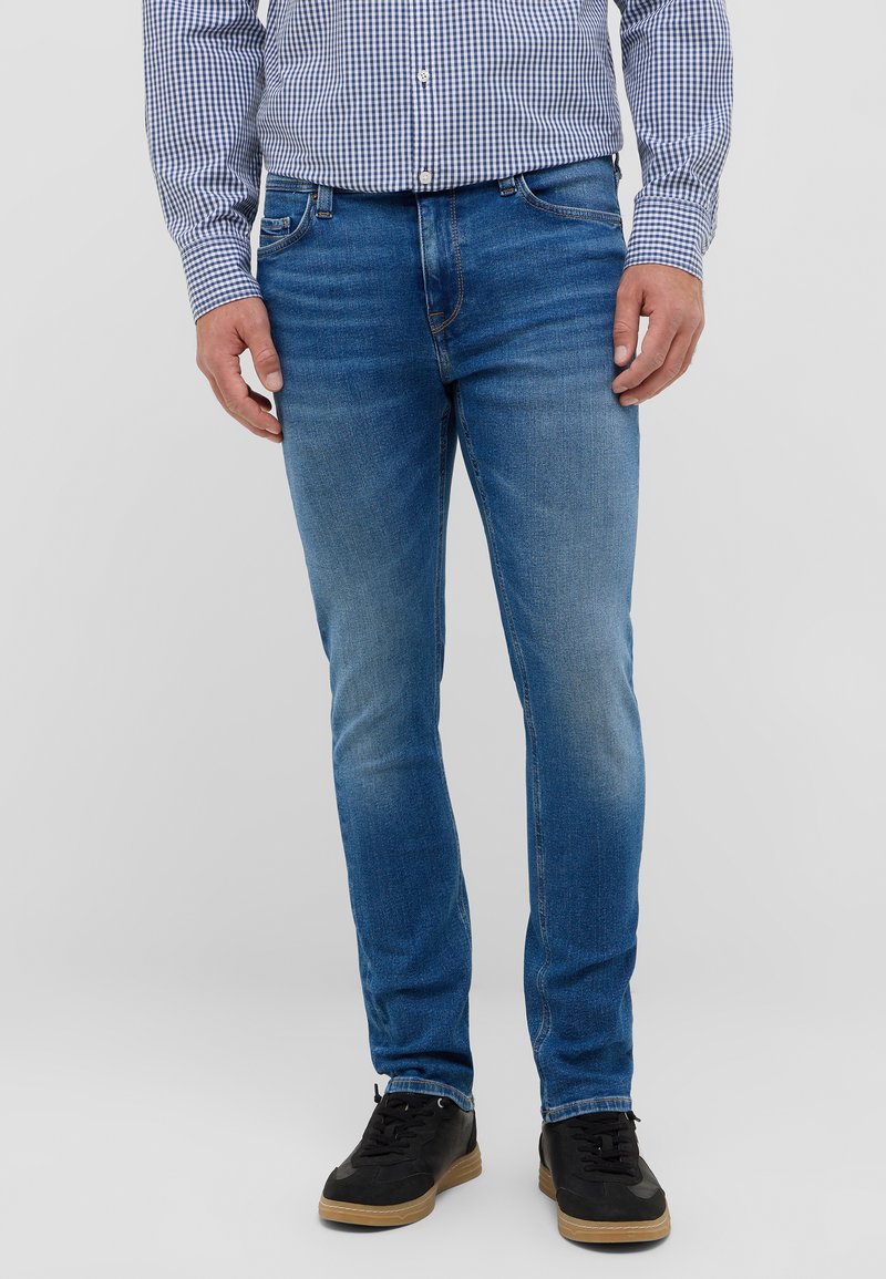 Niebieskie dżinsy, dopasowane, z wyblakłym wykończeniem. Noszone z koszulą w kratę i czarnymi sneakersami. Akcenty obejmują widoczne szwy.