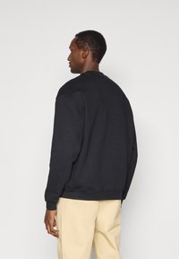 Αρσενικός στραμμένος αριστερά με κοντά μαλλιά, φορώντας ένα απλό μαύρο φούτερ και μπεζ παντελόνι μπροστά από έναν απλό ανοιχτό φόντο.