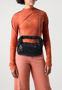 Svart paljettveske med avrundet topp, tykk hank og kjedehank, kombinert med en oransje langermet topp og rosa bukser.