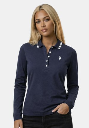 Woman with long blonde hair wearing a navy blue long-sleeve polo shirt with white collar stripes and a small white logo on the chest.