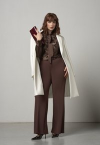 Pantaloni marroni a gamba larga abbinati a una blusa marrone con paillettes e fiocco, e un cappotto bianco. Porta una pochette rosso scuro e indossa tacchi neri.