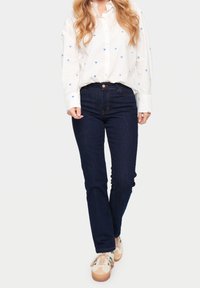Camicia bianca con motivi a cuore blu, jeans blu scuro e sneakers beige. La camicia ha maniche lunghe e un design con bottoni.