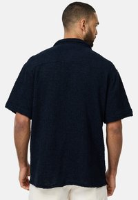 Camisa de manga corta azul marino hecha de tela texturizada. Presenta un corte holgado y un cuello, con costuras visibles y un dobladillo curvado.