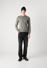 Groene gebreide trui met een klein logo, gecombineerd met zwarte rechte jeans en zwarte schoenen. Een zwarte beanie maakt de outfit compleet.