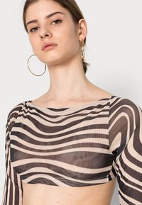 Top de mangas longas em tecido transparente, apresentando bold listras onduladas em preto e bege, com decote redondo e detalhes de costura visíveis.