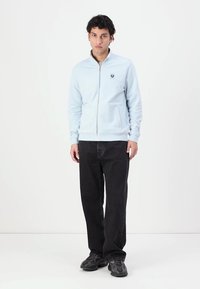 Jaqueta azul clara com fecho zip na frente e um pequeno logótipo no peito, usada sobre calças pretas de perna reta e ténis escuros. Textura de tecido suave.