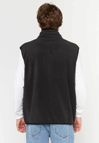 Chaleco de forro polar negro con cuello alto, sin mangas, textura suave y un ajuste relajado, llevado sobre una camiseta de manga larga blanca.