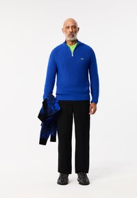 Blauwe geribde trui met een ritskraag, in laag over een groene polo. Zwarte cargobroek en zwarte sneakers, houdend een blauwe gebreide trui.