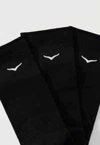 Τρεις μαύρες κάλτσες τοποθετημένες δίπλα η μία στην άλλη, η κάθε μία με ένα μικρό λευκό λογότυπο σε σχήμα πουλιού κοντά στη μανσέτα, σε απλό λευκό φόντο.