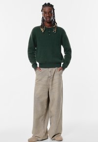 Zelený pletený sveter s golierom a troch gombíkoch, spárovaný s širokými béžovými džínsy a hnedou obuvou.