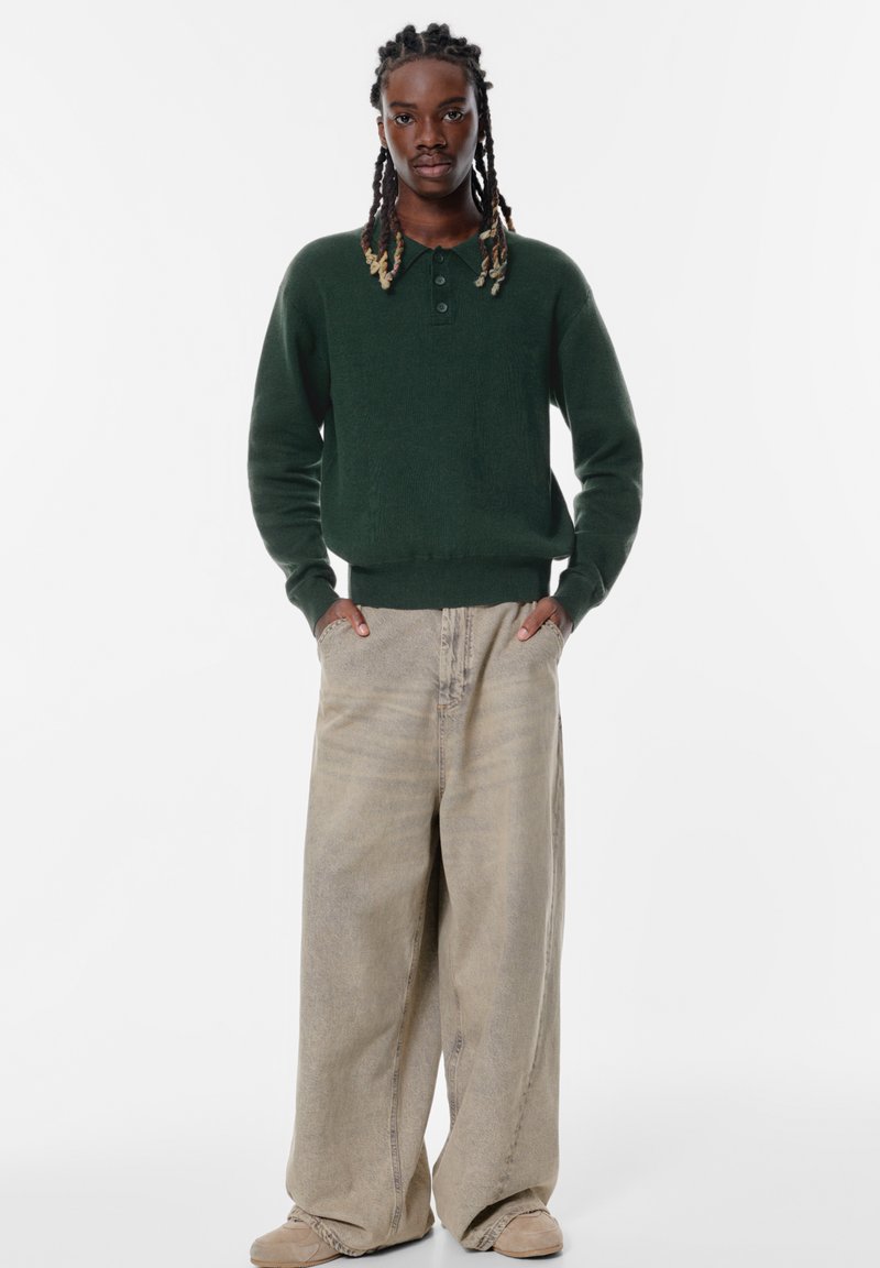 Zelený pletený sveter s golierom a troch gombíkoch, spárovaný s širokými béžovými džínsy a hnedou obuvou.