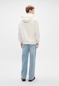 Ανοιχτό καφέ φούτερ με κουκούλα και άνετη εφαρμογή, εμφανίζεται από πίσω. Συνδυασμένο με ανοιχτό μπλε παντελόνι σε ίσια γραμμή και μαύρα παπούτσια. Λείος στίγμα.