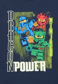 Dunkelblaues T-Shirt mit drei Comic-Ninja-Figuren in Blau, Grün und Rot, dazu auffälliger "DRAGON POWER"-Text in Weiß und grünem Farbspritz-Hintergrund.
