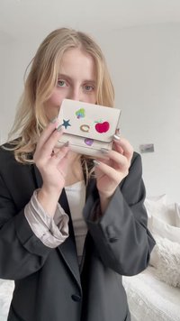 Portofel din piele albă, decorat cu autocolante colorate, inclusiv o stea, un măr, o inimă și un copac. Dispune de un închizător auriu și are o formă rectangulară compactă.