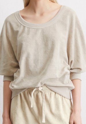 Femme portant un haut beige à manches courtes ample avec un col côtelé et un pantalon à cordon assorti, sur un fond uni.