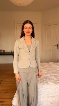 Νεαρή γυναίκα που φοράει ανοιχτό γκρι κοστούμι με μπλέιζερ και φαρδιά παντελόνια, στέκεται σε υπνοδωμάτιο με ξύλινο πάτωμα και λευκά σεντόνια.
