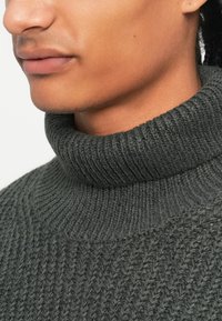 Mørkegrå sweater med rullekrave og en struktureret strikket mønster, der har en ribbet krave og en glat krop, hvilket giver et hyggeligt udseende.