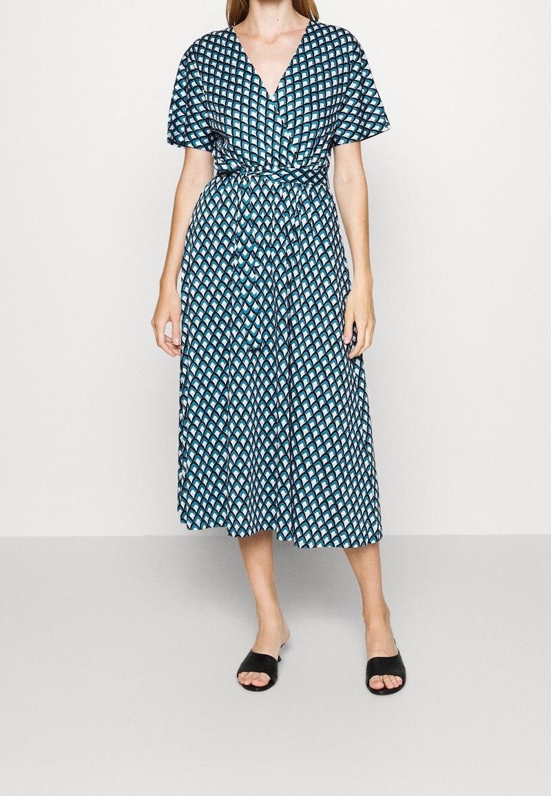 Femme portant une robe midi bleue à motifs avec des manches courtes, un col en V cache-cœur et une ceinture nouée à la taille, associée à des chaussures noires sans lacets.