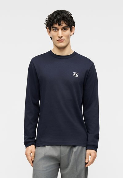 Νέος άνδρας με σγουρά σκούρα μαλλιά φοράει μπλε σκούρο πουκάμισο μακρυμάνικο με λογότυπο στο στήθος και γκρι παντελόνια, στέκεται μπροστά σε απλό φόντο.