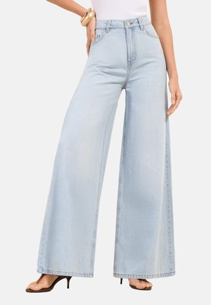 Kvinde iført lyseblå jeans med høj talje og vide ben, hvid ærmeløs top, guldbøjle og sorte åben-tå hælede sandaler.