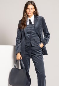 Granatowy garnitur z korduroyu z dopasowaną marynarką, kamizelką zapinaną na pięć guzików oraz prostymi spodniami. Połączony ze sztywną torebką w dopasowanym odcieniu.