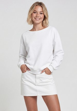 Ung kvinde med kort blondt hår iført en hvid langærmet top og hvidt skørt, smilende med hænderne i lommerne foran en ensfarvet baggrund.
