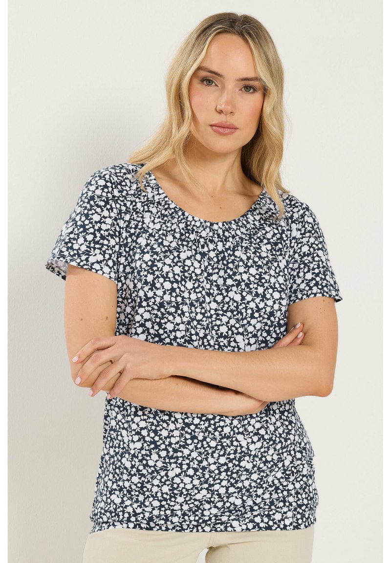 Femme aux cheveux blonds portant un chemisier à manches courtes noir et blanc à fleurs, bras croisés, debout devant un fond clair uni.