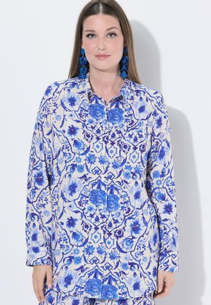 Kobieta ubrana w niebiesko-białą koszulę zapinaną na guziki w kwiatowy wzór oraz pasujące kolczyki, stojąca na tle jednolitego białego tła.