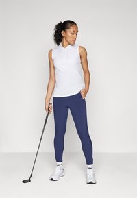 Valkoinen hihaton polo-paita ja tummansiniset legginsit, yhdistettynä valkoisiin urheilukenkiin. Pitää mustaa golf-mailaa. Sujuva kangas.