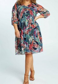 Nainen, jolla on yllään värikäs, polvet peittävä mekko trooppisella lehväkuviolla ja kolmivartiset hihat, yhdistettynä vaaleisiin nilkkasandaaleihin.