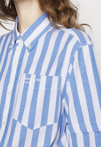 Camicia a righe blu e bianche realizzata in tessuto leggero, con colletto a bottoni, taschino sul petto e piccoli dettagli con logo.