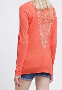 Coral long-sleeve shirt with intricate lace detail in a V-shape at the back, featuring a relaxed fit and soft fabric texture.