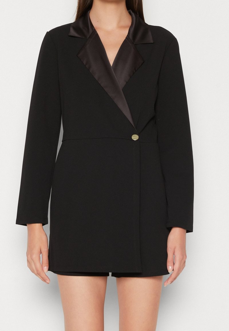 Femme portant une robe blazer noire avec des revers en satin et un bouton doré unique, debout devant un fond blanc uni.