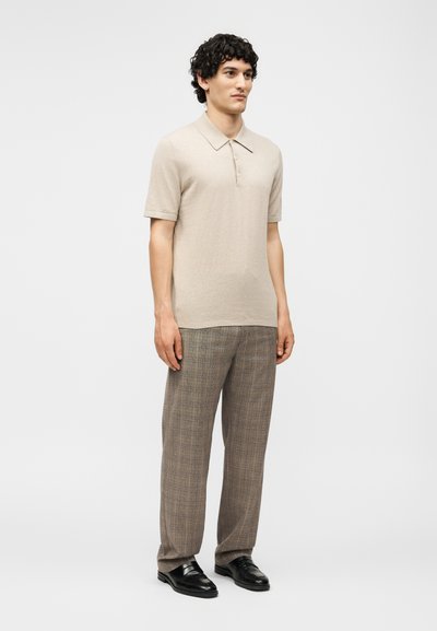 Άνδρας που στέκεται φορώντας ένα μπεζ πόλο με κοντά μανίκια, καφέ καρό παντελόνια και μαύρα δερμάτινα παπούτσια σε απλό φόντο.