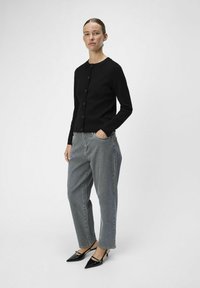 Cardigan noir à boutons avec des manches longues, associé à un jean taille haute gris. Le modèle porte des escarpins noirs brillants à bout pointu.