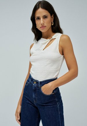 Top sin mangas texturizado en blanco con un detalle de escote diagonal y un acento dorado, combinado con unos jeans de cintura alta azul oscuro.