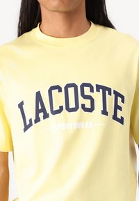 Geltona medvilninė marškinėlė su ryškiu tamsiai mėlynu „LACOSTE“ logotipu ir baltu tekstu „SPORTSWEAR“ žemiau. Standartinė apvalios apykaklės, trumpomis rankovėmis.