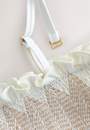 Weißer strukturierter Stoff mit gerafftem Rand und beiger Naht, ausgestattet mit verstellbaren weißen Trägern mit goldfarbenen Haken und Ringen.