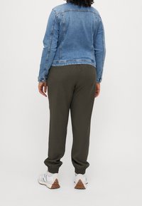 Person som står vänd bort med ryggen mot kameran, iklädd en blå jeansjacka, mörkgröna joggerbyxor, vita strumpor och vita sneakers med bruna sulor.