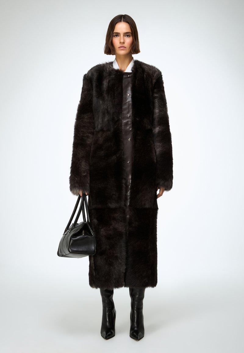 Long manteau en fausse fourrure foncée avec un col, associé à une robe en cuir sombre et un sac à main noir, porté avec des bottes montant aux genoux.