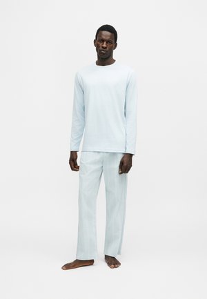 Homme debout pieds nus portant une chemise à manches longues bleu clair et un pantalon ample assorti sur un fond blanc uni.