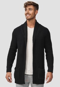 Cárdigan negro de punto con cuello de chal, dos bolsillos frontales, puños acanalados y tejido texturizado. Llevado sobre una camisa de color claro.