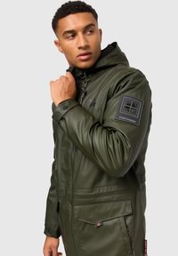 Green waterproof jacket with a hood, featuring a zip closure, multiple pockets, and a logo badge on the sleeve. Smooth, shiny texture.