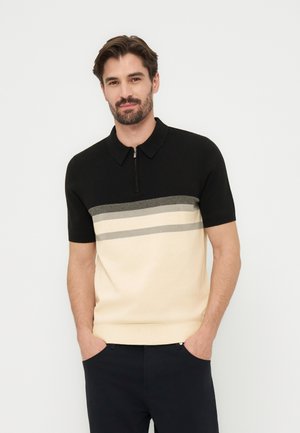 Homme barbu portant un polo à manches courtes noir, gris et beige avec une fermeture éclair, debout les mains dans les poches devant un fond blanc uni.