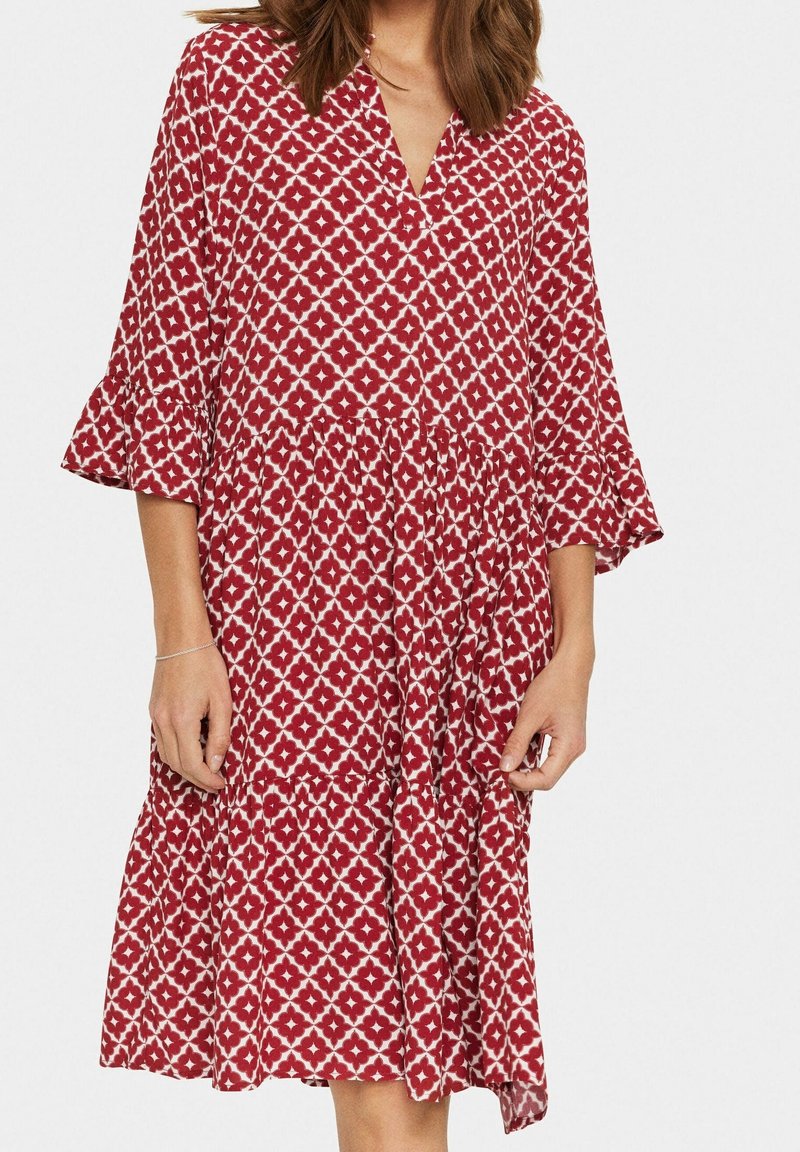 Femme vêtue d'une robe mi-genoux à motif rouge et blanc avec des manches évasées trois-quarts et un décolleté en V.