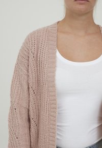 Stickad cardigan i mjukt rosa, med en lös passform, ribbade ärmslut och intrikata flätningsmönster. Bärs över en vit linne.