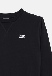 Czarna bluza z ribbowanym dekoltem w serek, z białym haftowanym logo New Balance na lewym piersie. Miękka tkanina, minimalistyczny design.