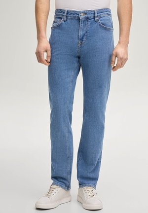 Mężczyzna w jasnoniebieskich dopasowanych dżinsach i białych trampkach, stojący na tle jednolitego jasnego tła.